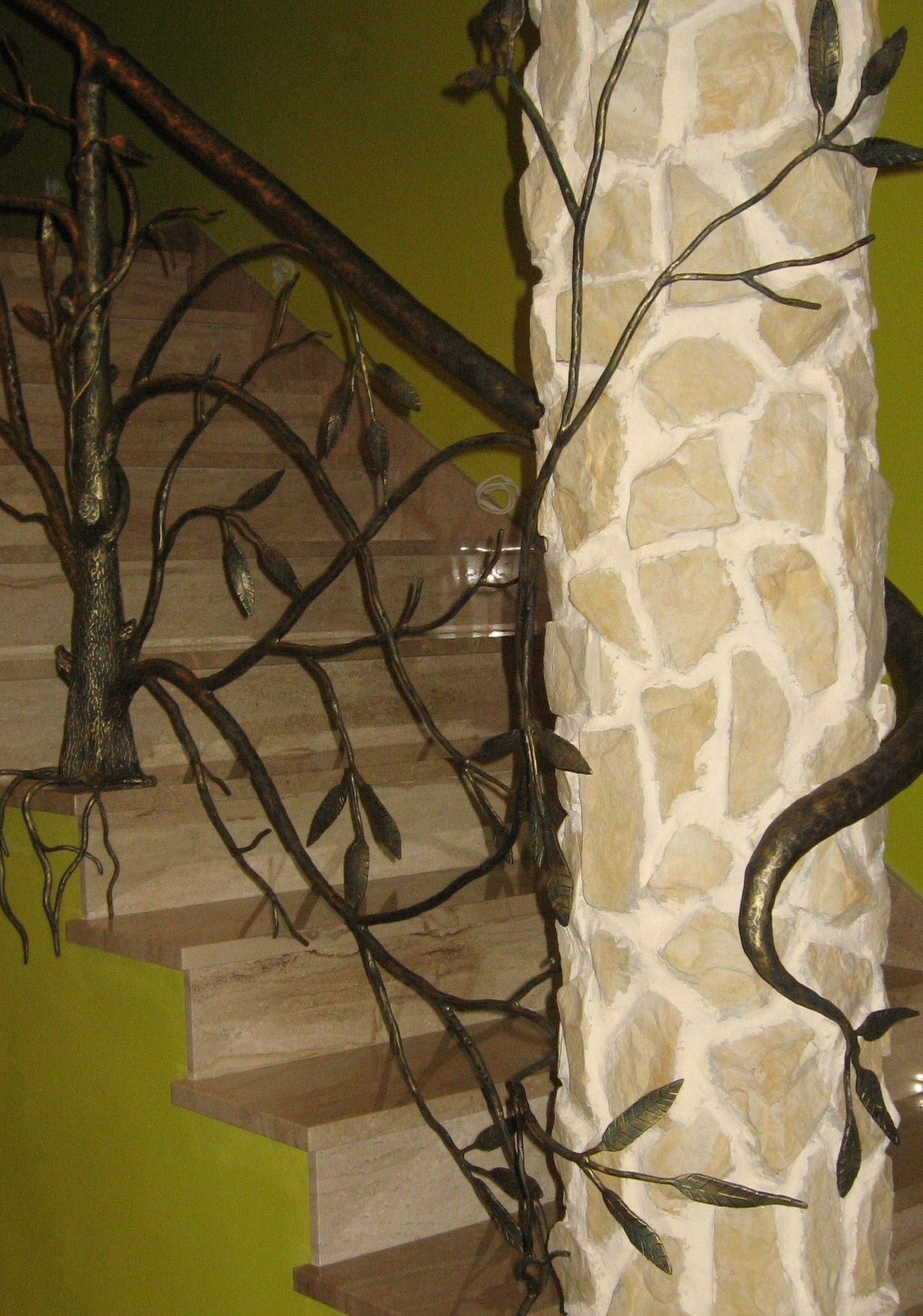 Kute balustrady schodowe w formie drzewa z liśćmi, oplatające kamienny filar, na tle zielonej ściany i marmurowych schodów.